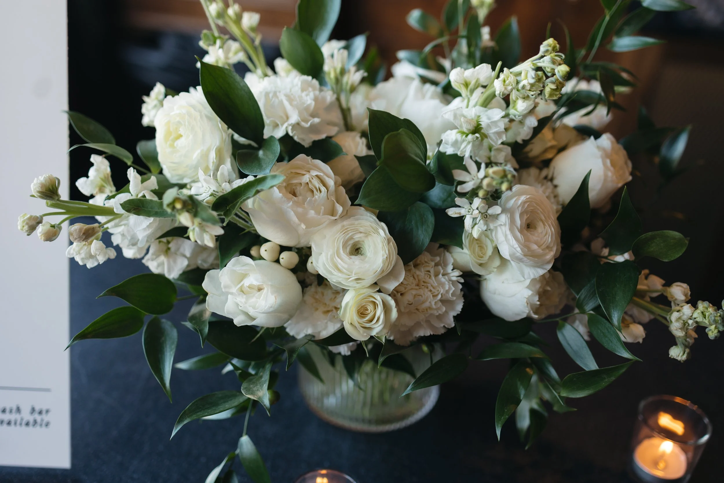 A bouquet of white flowers including roses and other blooms with green leaves, arranged on a dark surface with small tealight candles nearby.