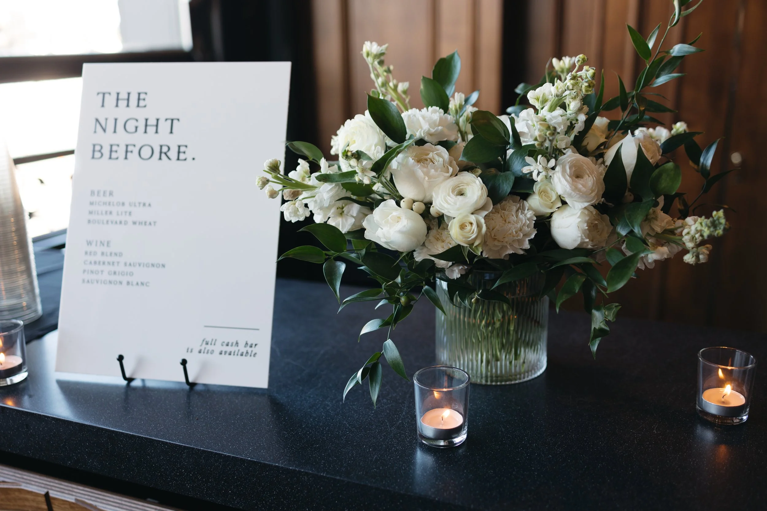 A large clear glass vase filled with white flowers and green leaves, placed on a black table. Next to it is a white card with black text reading "THE NIGHT BEFORE." and a list of wine options. Surrounding the vase are small lit candles in glass holde