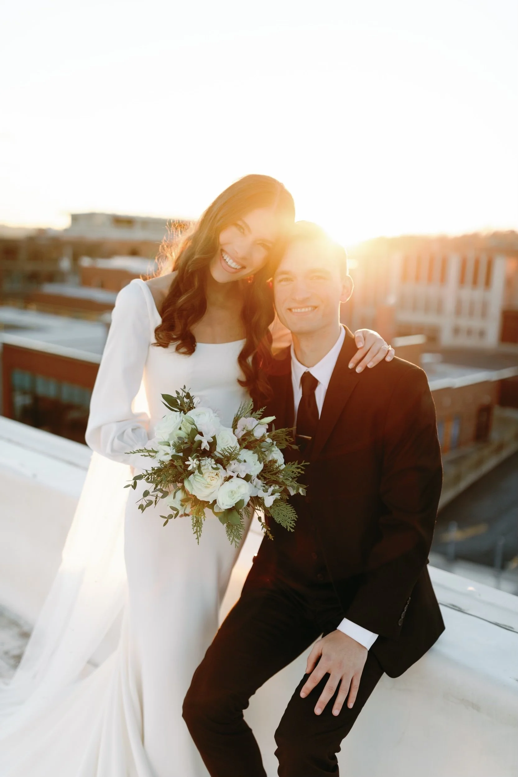 A smiling woman and man dressed in wedding attire, standing on a rooftop during sunset, with the woman holding a bouquet of white flowers and greenery.