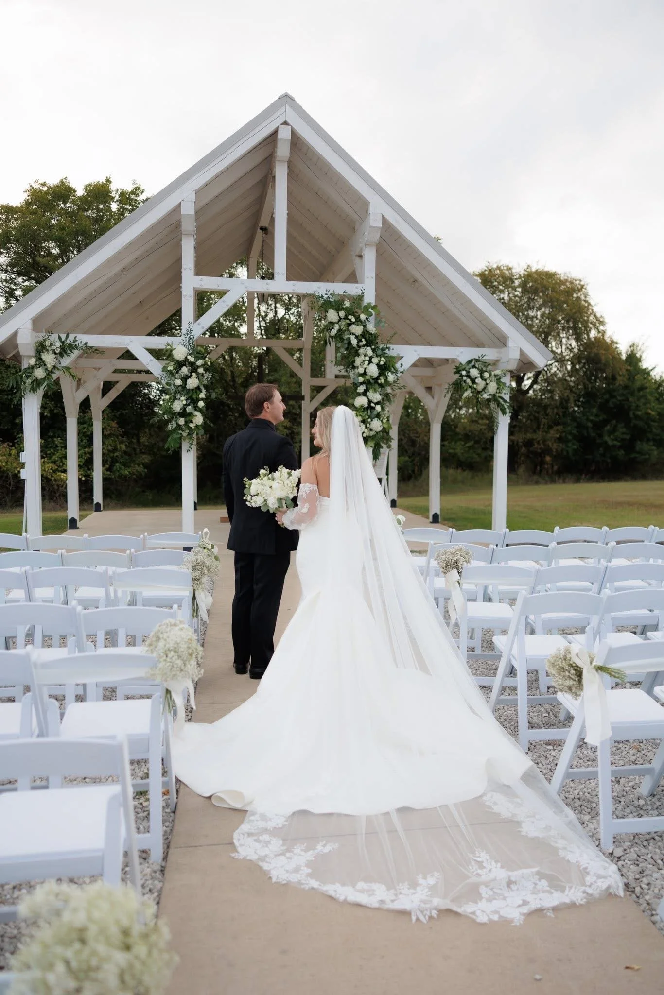 A bride and groom standing under a wooden outdoor wedding arch decorated with white flowers, during their wedding ceremony in a garden setting.