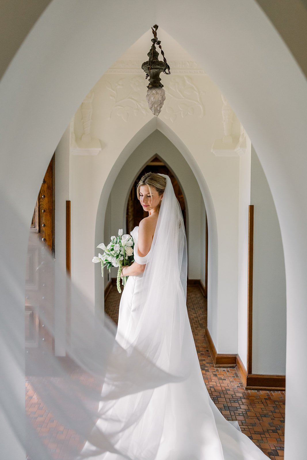 Bride in white wedding gown and veil holding a bouquet of white flowers, standing in a hallway with arched doorways.