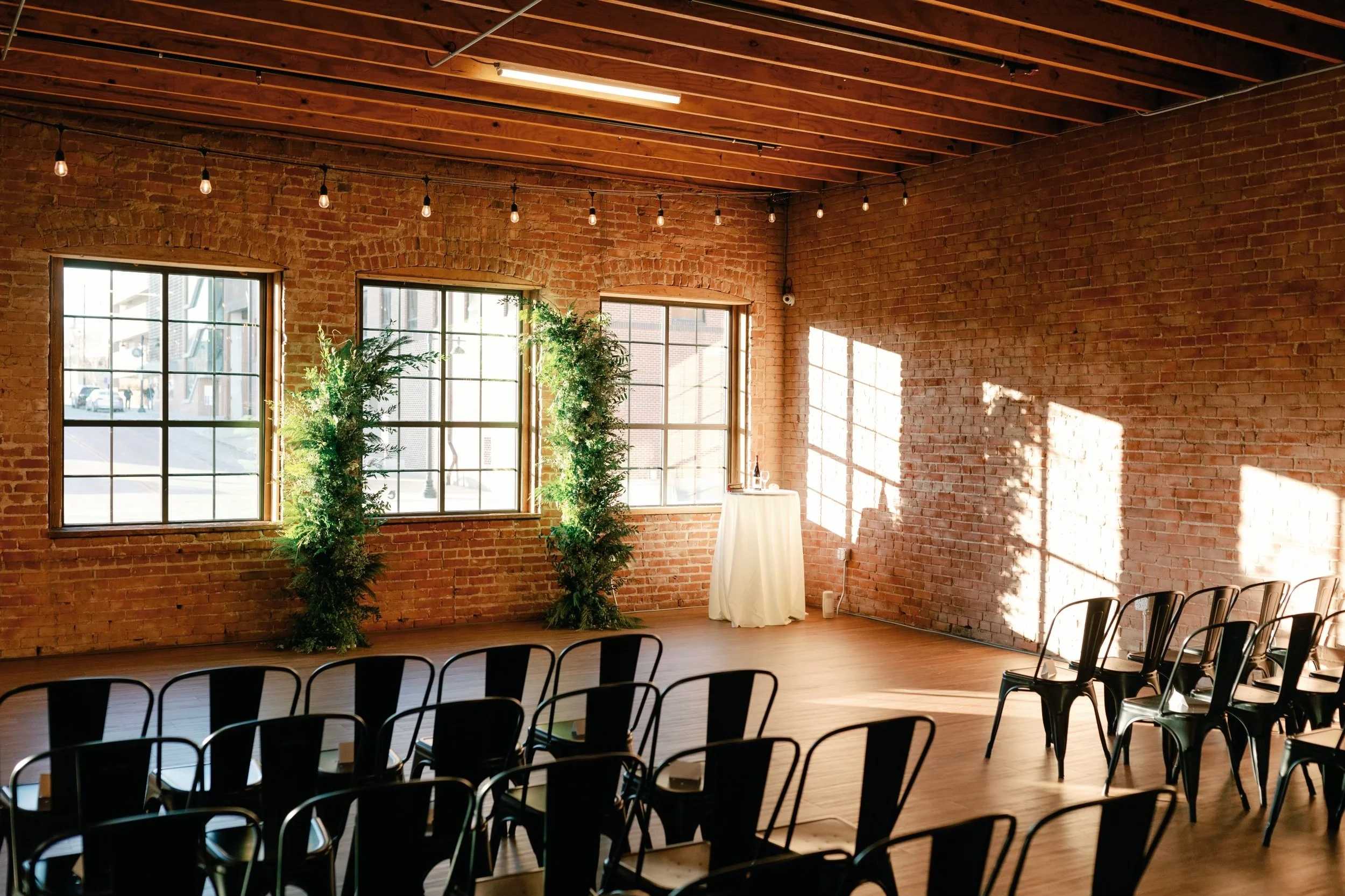 Indoor event space with exposed brick walls, large windows, and wooden ceiling beams. There are rows of black chairs arranged for an audience, and a small table with a white tablecloth near the windows. String lights hang from the ceiling.
