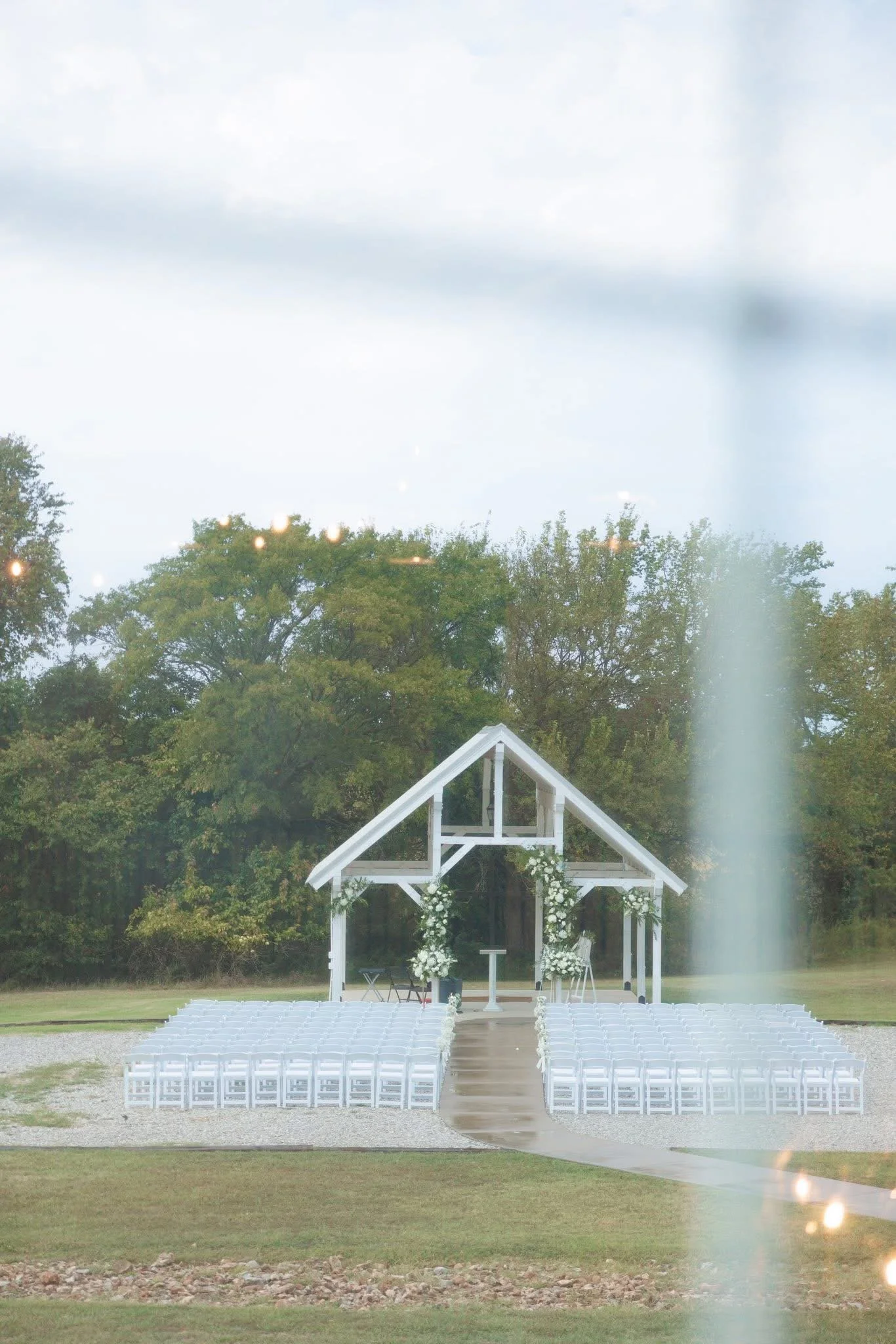 Outdoor wedding ceremony setup with white chairs arranged in rows on gravel, a white wooden arch decorated with white flowers, and a natural backdrop of trees under a cloudy sky.