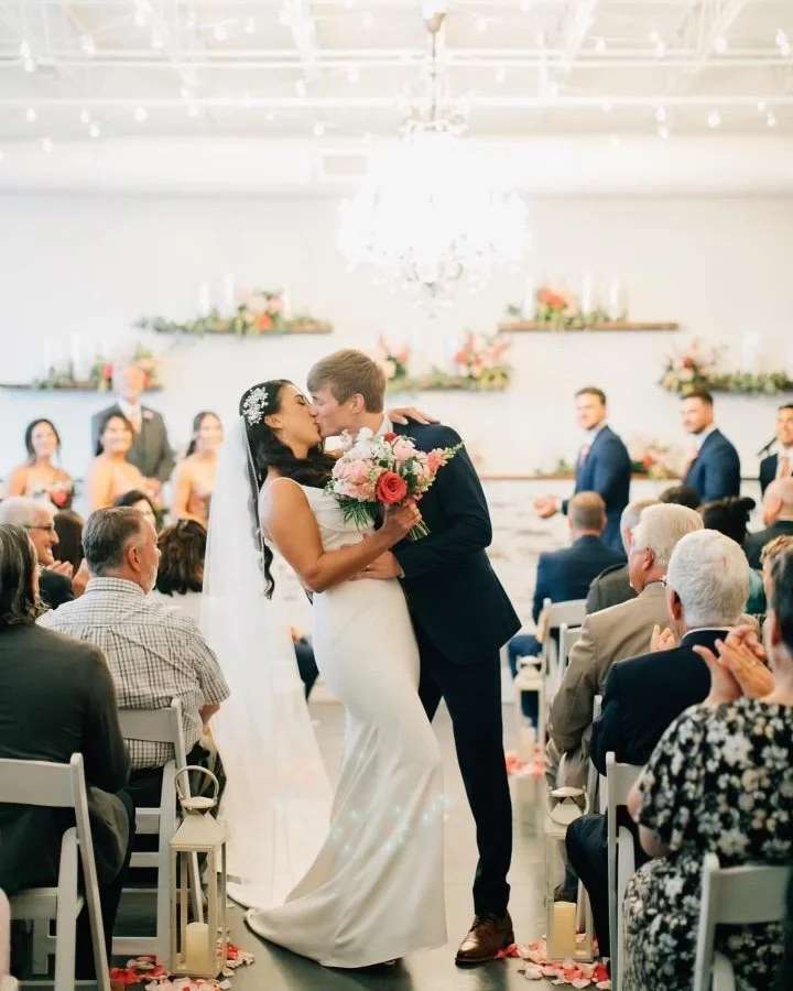 A bride and groom sharing their first kiss during their wedding ceremony in a bright, decorated venue, with guests seated on either side and bridesmaids and groomsmen in the background.