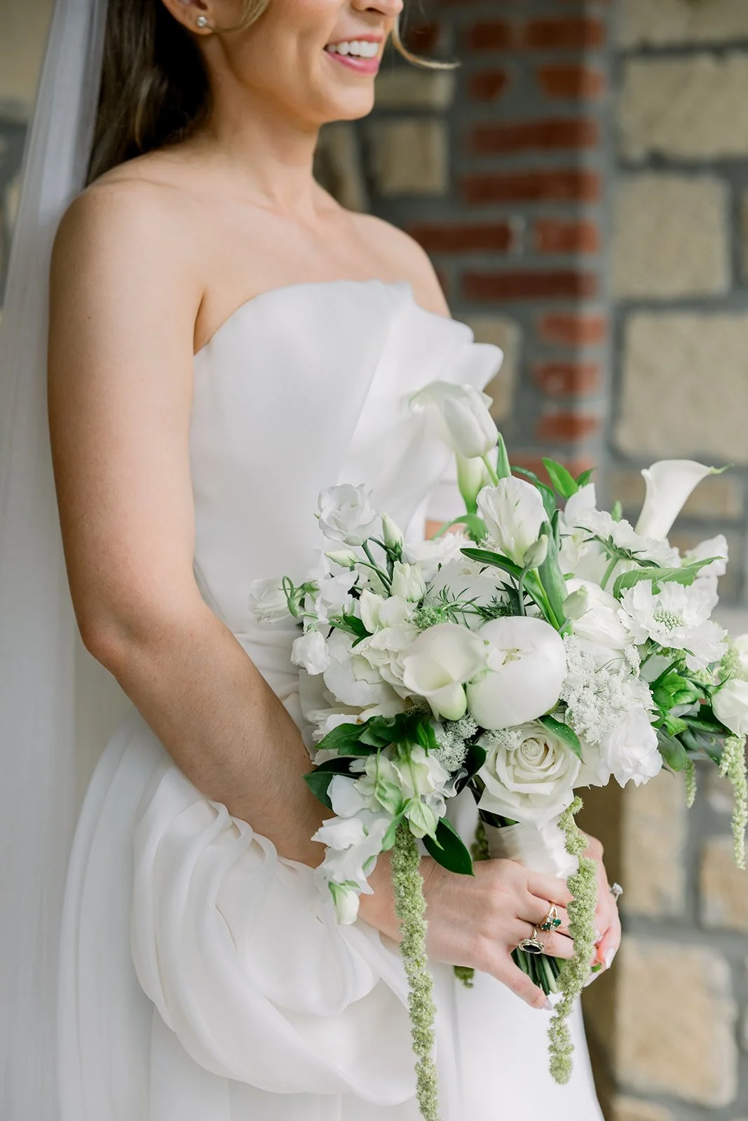 Close-up of a bride in a strapless white wedding gown holding a bouquet of white flowers in front of a brick wall.