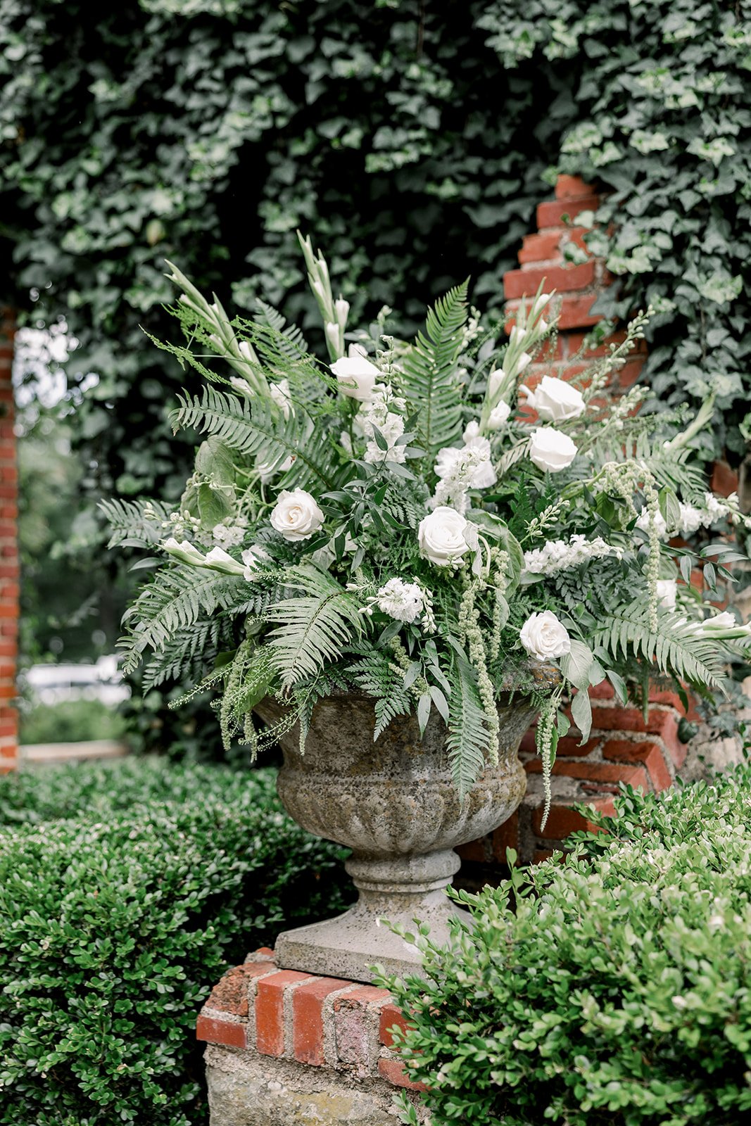 A large decorative stone planter filled with white roses, ferns, and other green foliage, set against a brick wall and lush greenery.