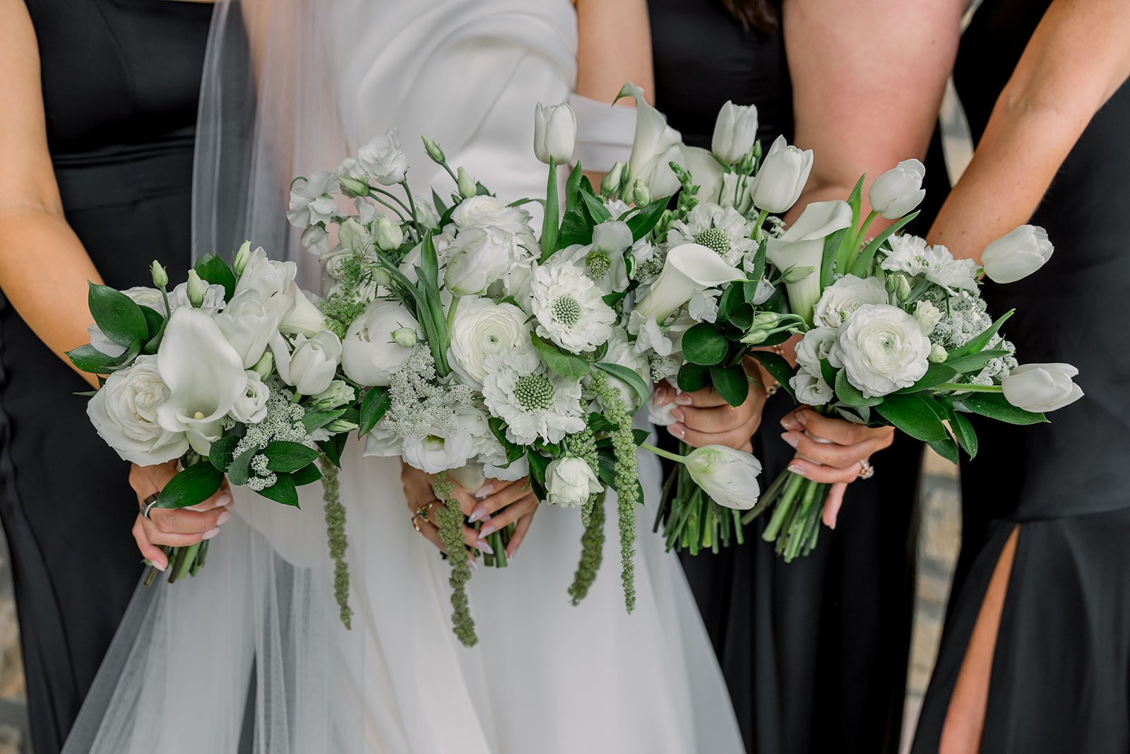 Three women holding bridal bouquets of white flowers, including tulips, roses, and greenery, at a wedding.