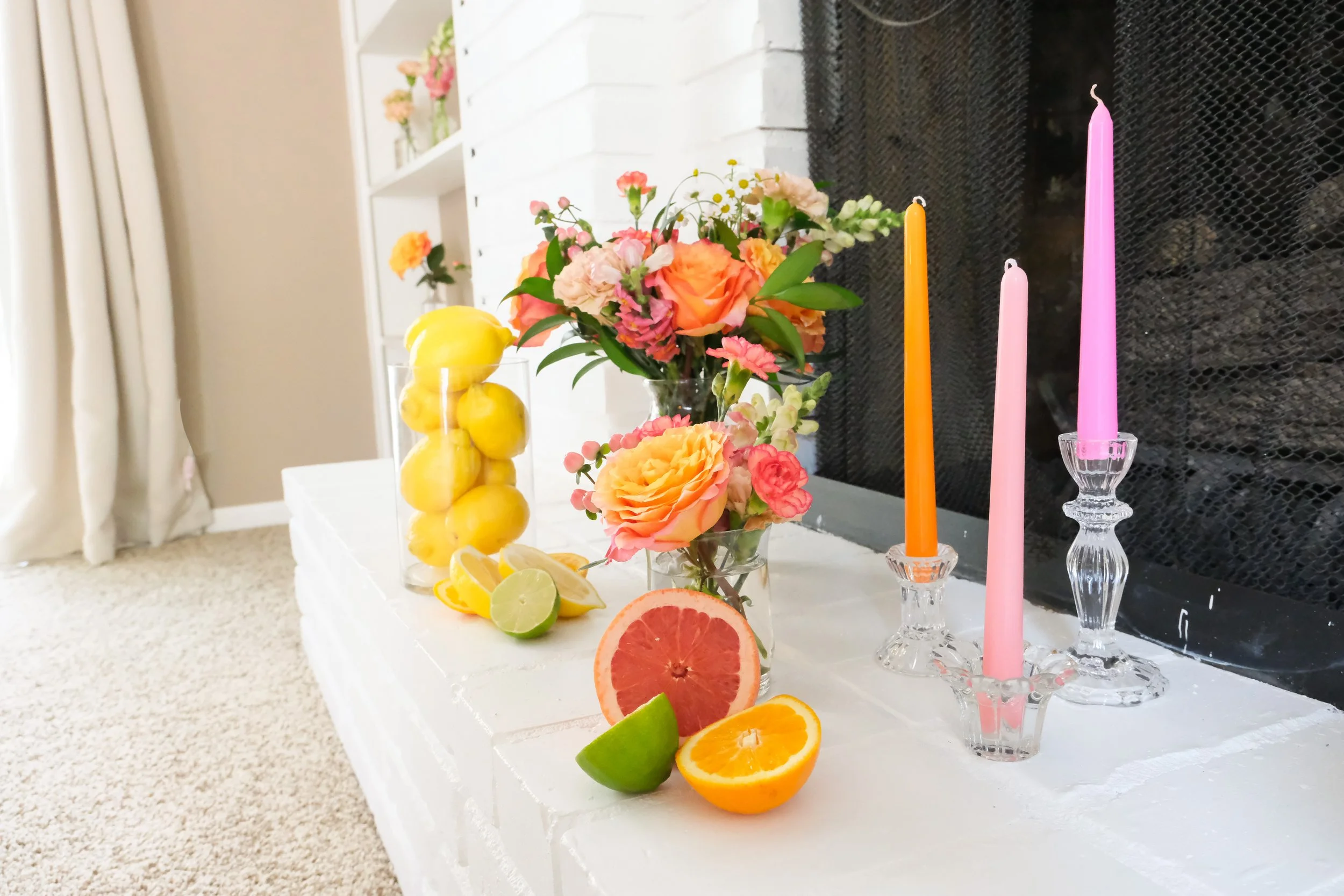 Arrangement of lemons, limes, oranges, and grapefruit slices with a bouquet of pink and orange flowers, pink and orange candles on a white table.