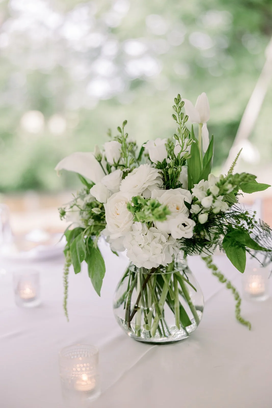 A clear glass vase filled with white flowers, including roses, tulips, and greenery, on a white tablecloth with blurred candles in the background.