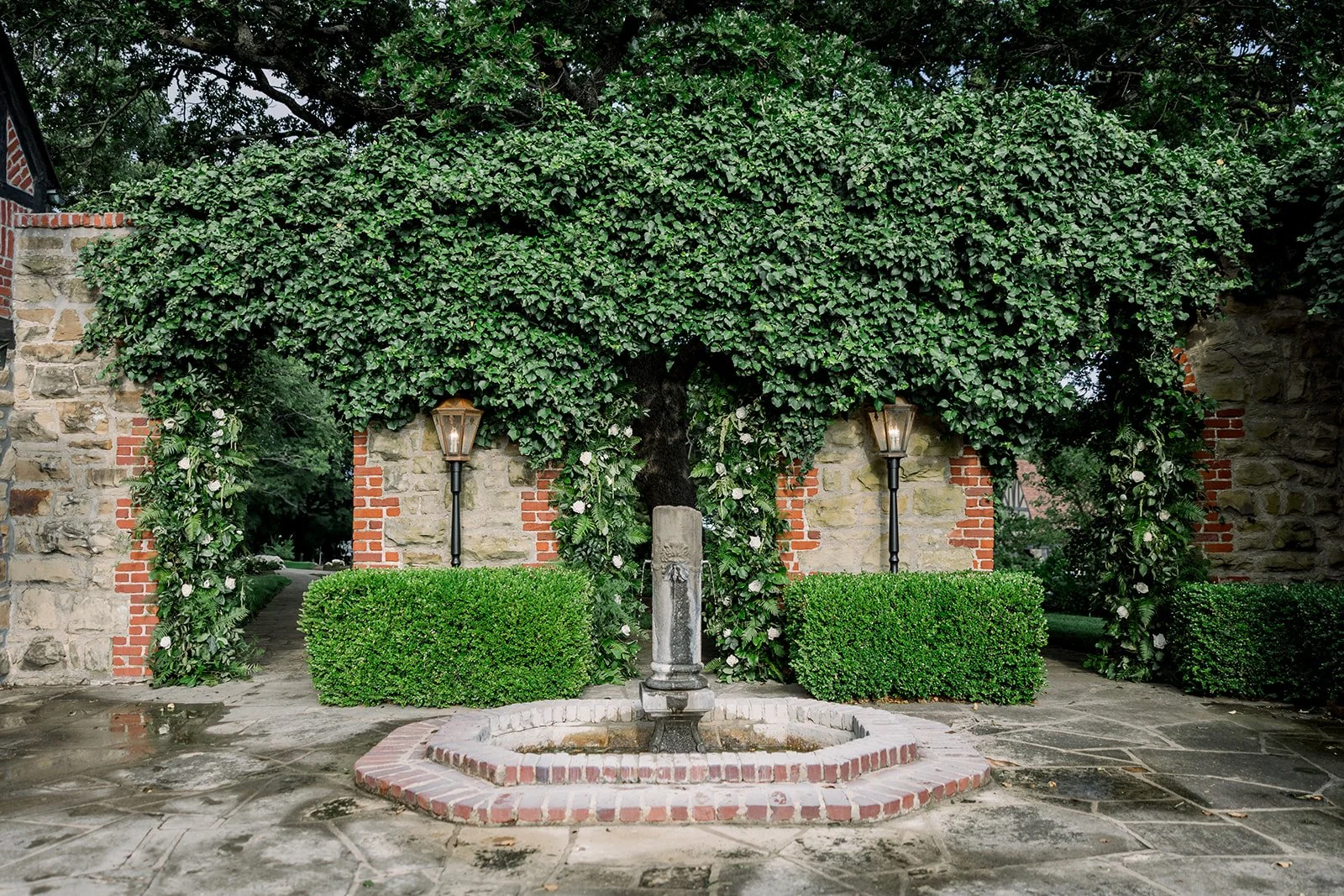 A decorative outdoor garden scene with a water fountain, two green trimmed bushes, two vintage-style lamps, an arch covered in green ivy, stone and brick walls, and a stone pathway.