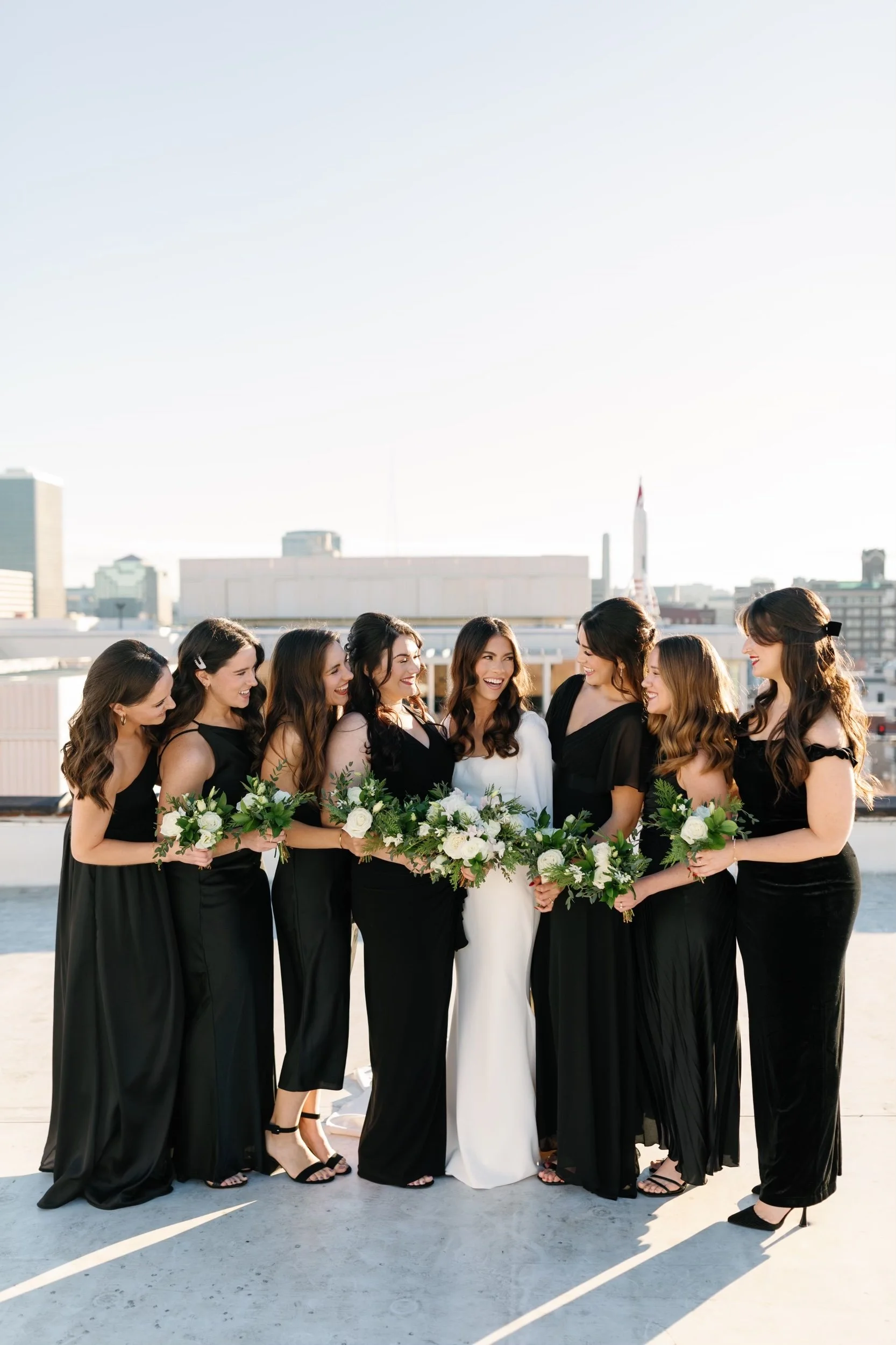 A group of women in formal black dresses and high heels standing on a rooftop, smiling and holding white flowers, with a city skyline in the background during the daytime.