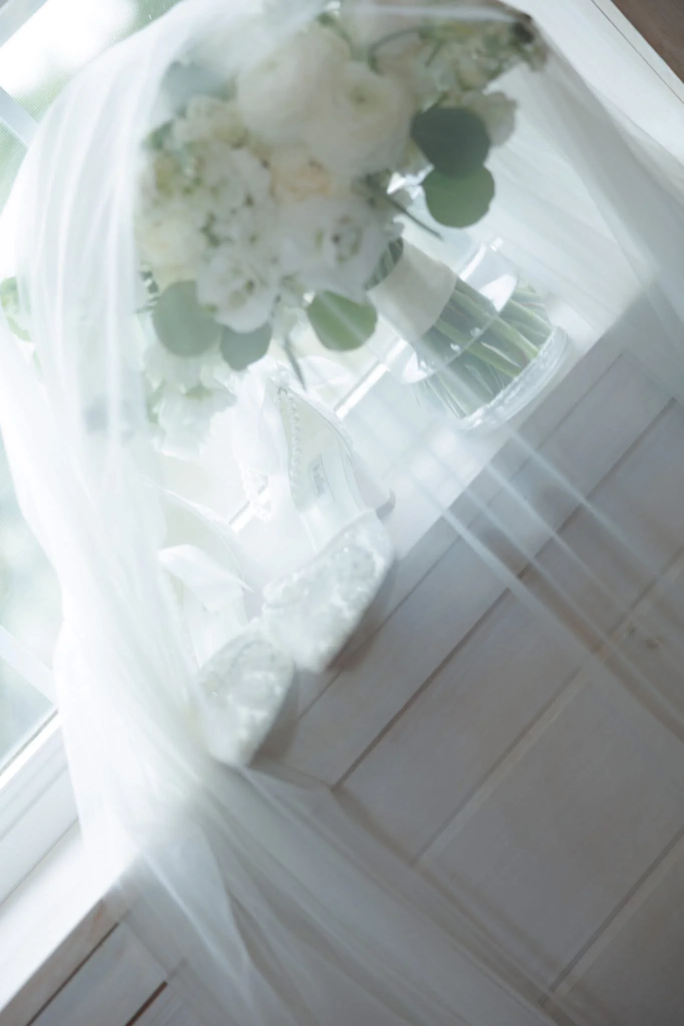 A bouquet of white flowers and greenery in a clear glass vase, placed on a windowsill with white horizontal panels.