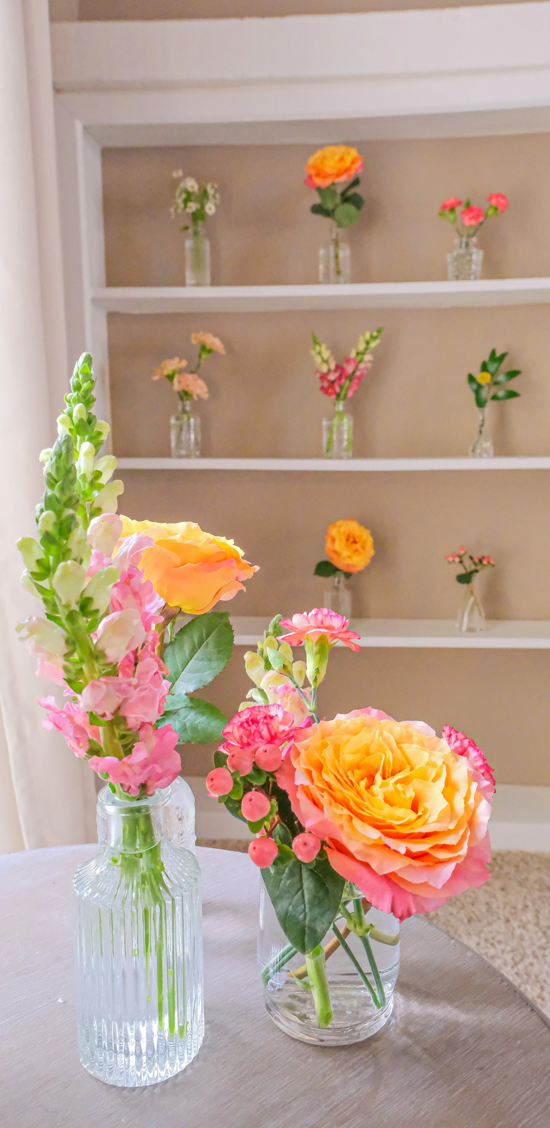 Vases with colorful flowers on a table and shelves in the background with similar floral arrangements.