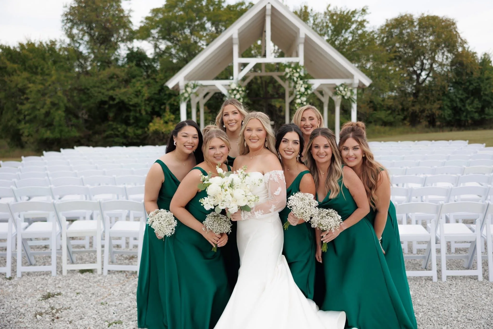 A bride in a white wedding gown holding a bouquet stands with six bridesmaids in green dresses during an outdoor wedding ceremony with white chairs and a white arch decorated with flowers, set against trees and an overcast sky.