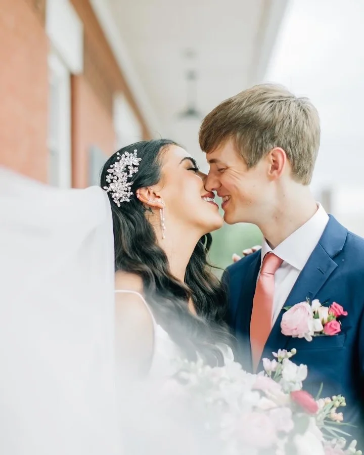 A bride and groom are about to kiss, standing close, with their noses touching, outdoors near a building, in a wedding setting.