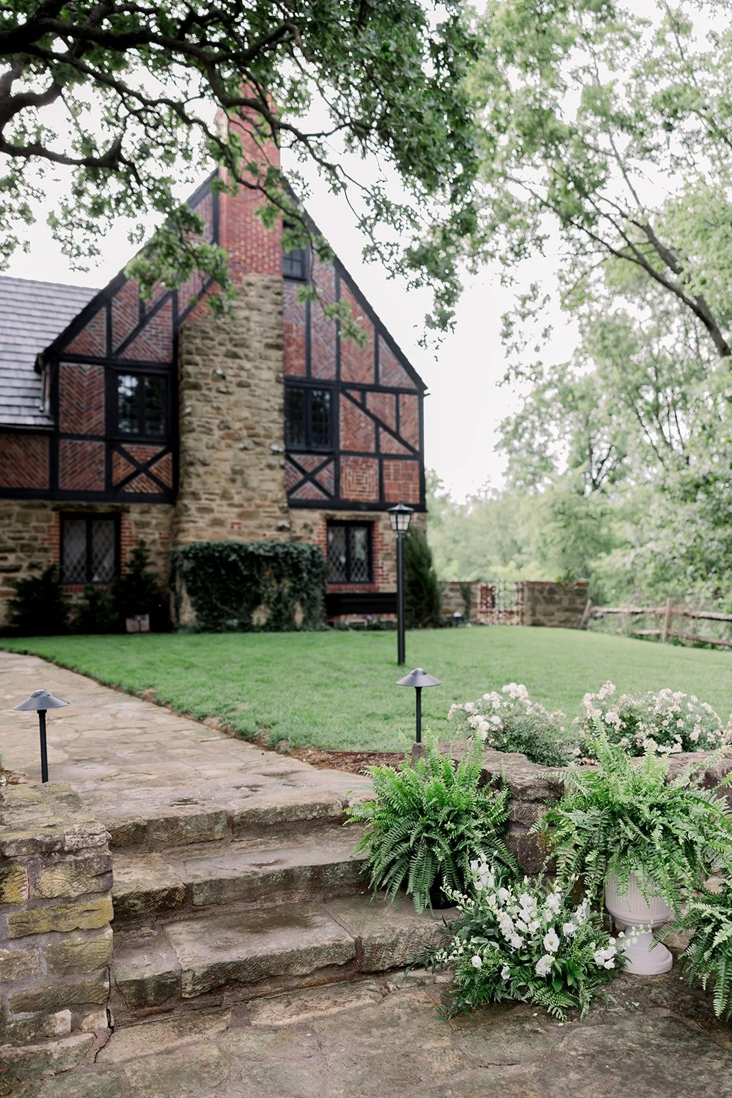 A stone and brick house with a chimney, surrounded by a green lawn, trees, and plants, with stone steps leading up to the house and outdoor lamps.