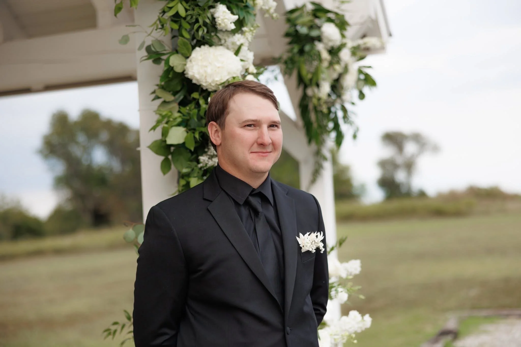 A man in a black suit standing outdoors beside a floral wedding arch with white flowers and greenery, with trees and an overcast sky in the background.