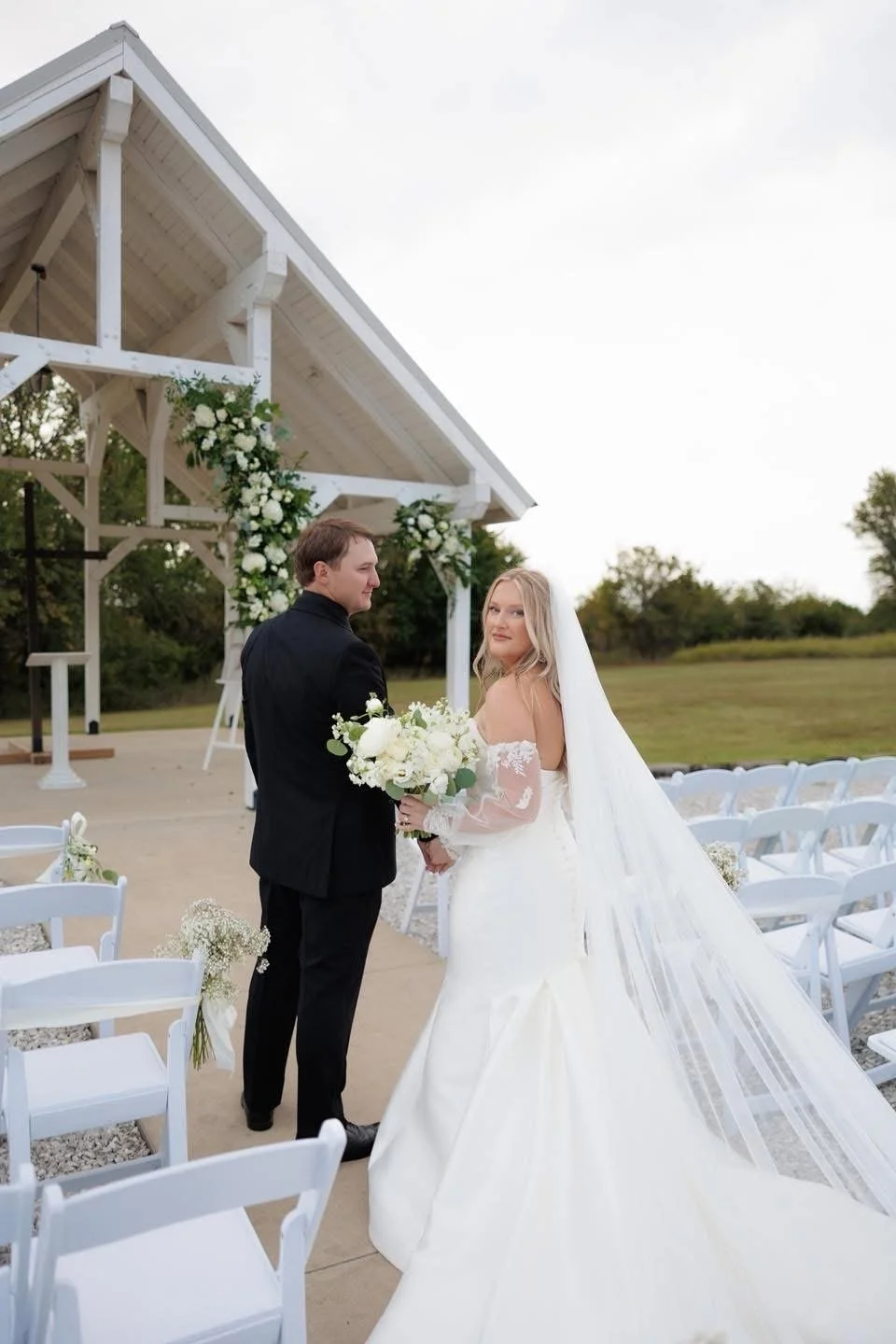 A bride and groom holding hands at an outdoor wedding ceremony, with chairs decorated with flowers, a white wooden arch with floral arrangements, and a grassy landscape in the background.