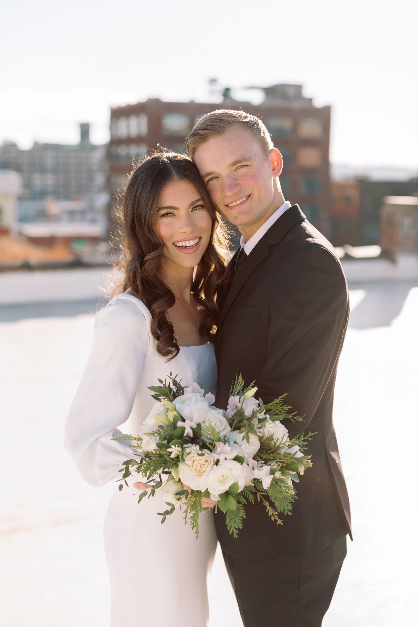 A bride and groom smiling and hugging each other outdoors with a cityscape in the background. The bride is holding a bouquet of white flowers and greenery. The bride has long, wavy brown hair and is wearing a white dress. The groom has short blond ha