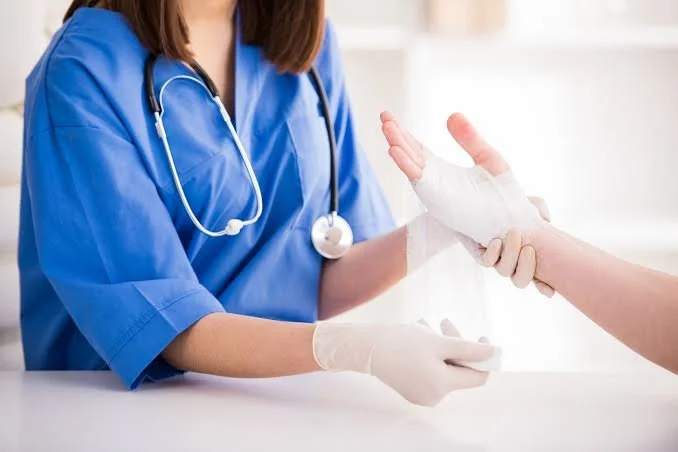 A healthcare professional in blue scrubs and a stethoscope assists a patient with a bandaged hand at a medical facility.