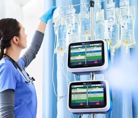 A nurse in blue scrubs and gloves setting up IV bags in a hospital with two monitors displaying patient information