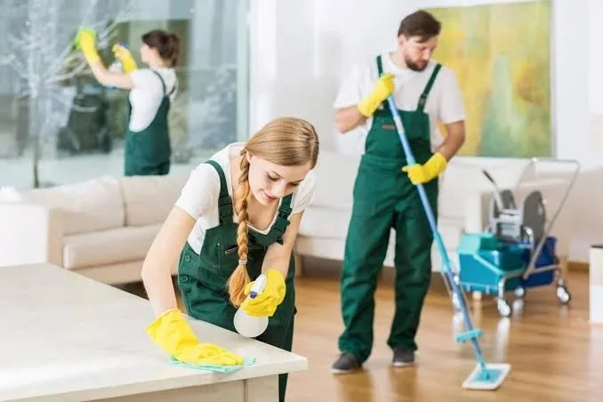 Three people cleaning a living room, wearing green uniforms and yellow gloves, with cleaning supplies.