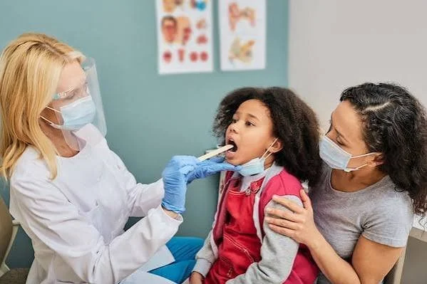 A healthcare professional in protective gear conducts a throat examination on a young girl in a medical clinic.