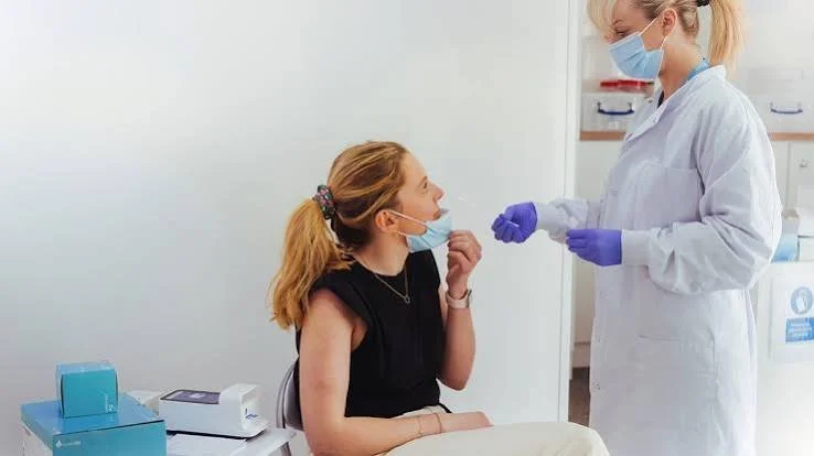 A healthcare professional doctor or nurse wearing a face mask and gloves is preparing to administer a COVID-19 vaccine or test to a woman sitting in a clinic, both wearing face masks.