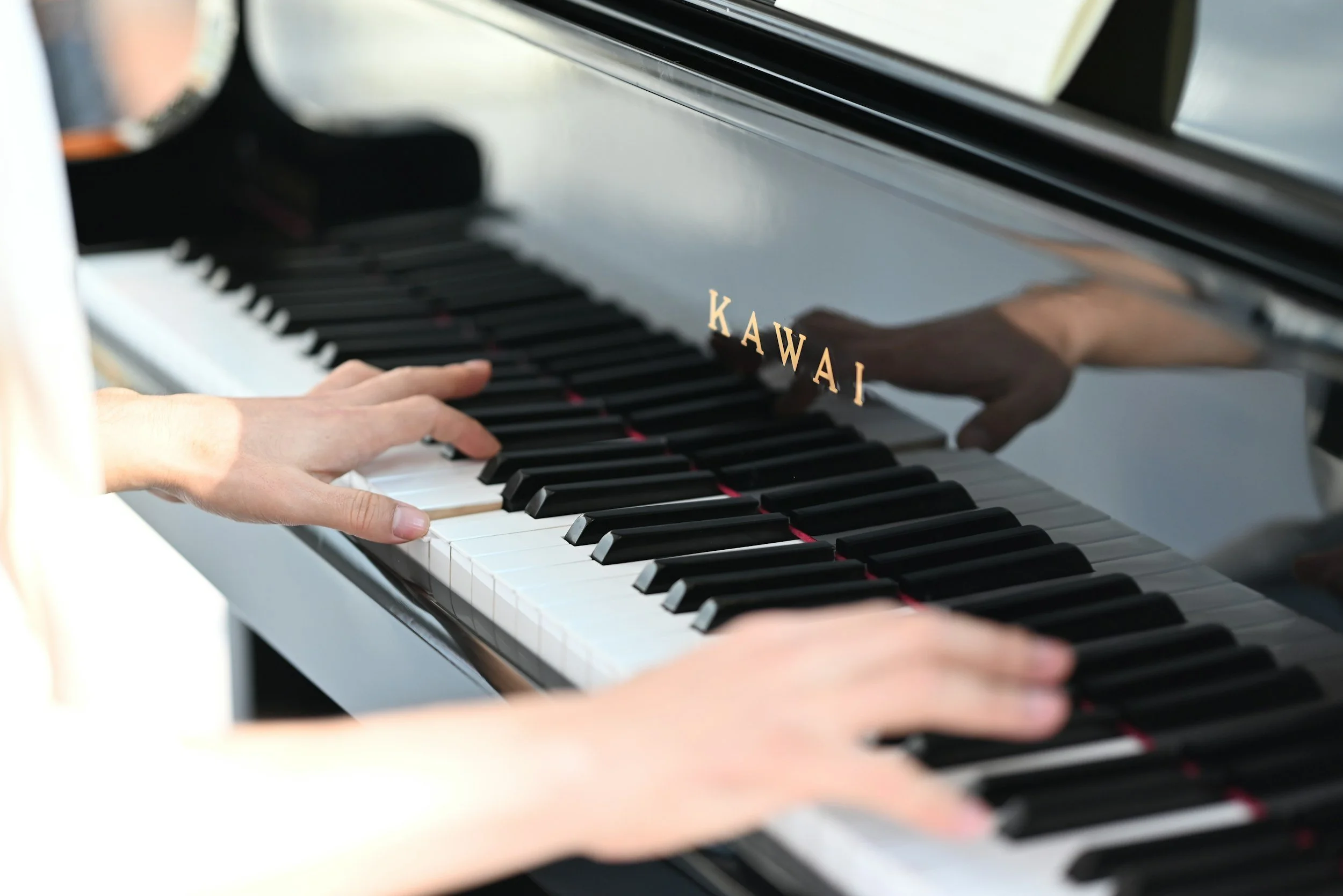 Close-up of hands playing a black grand piano with the brand name 'KAWAI' in gold lettering on the side, reflecting in the polished surface.