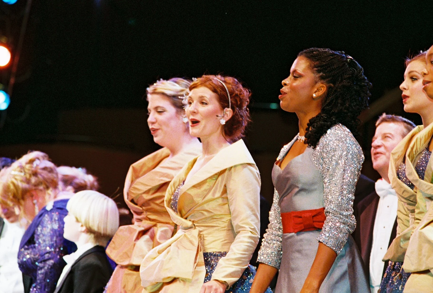 Women and children dressed in colorful costumes, singing or performing on stage with dark background and stage lights.