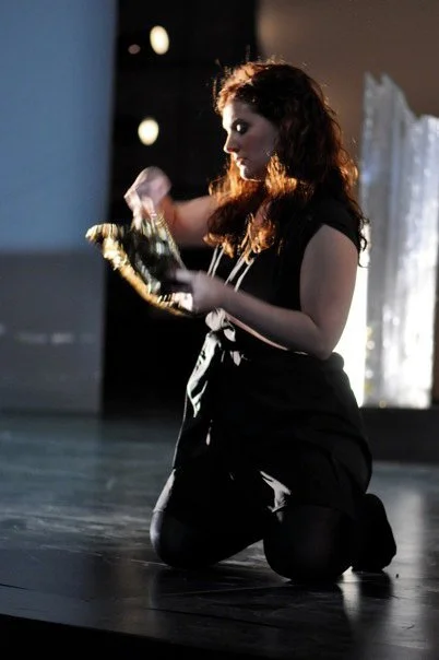 A woman with curly hair kneeling on stage, holding a shoe and a purse.