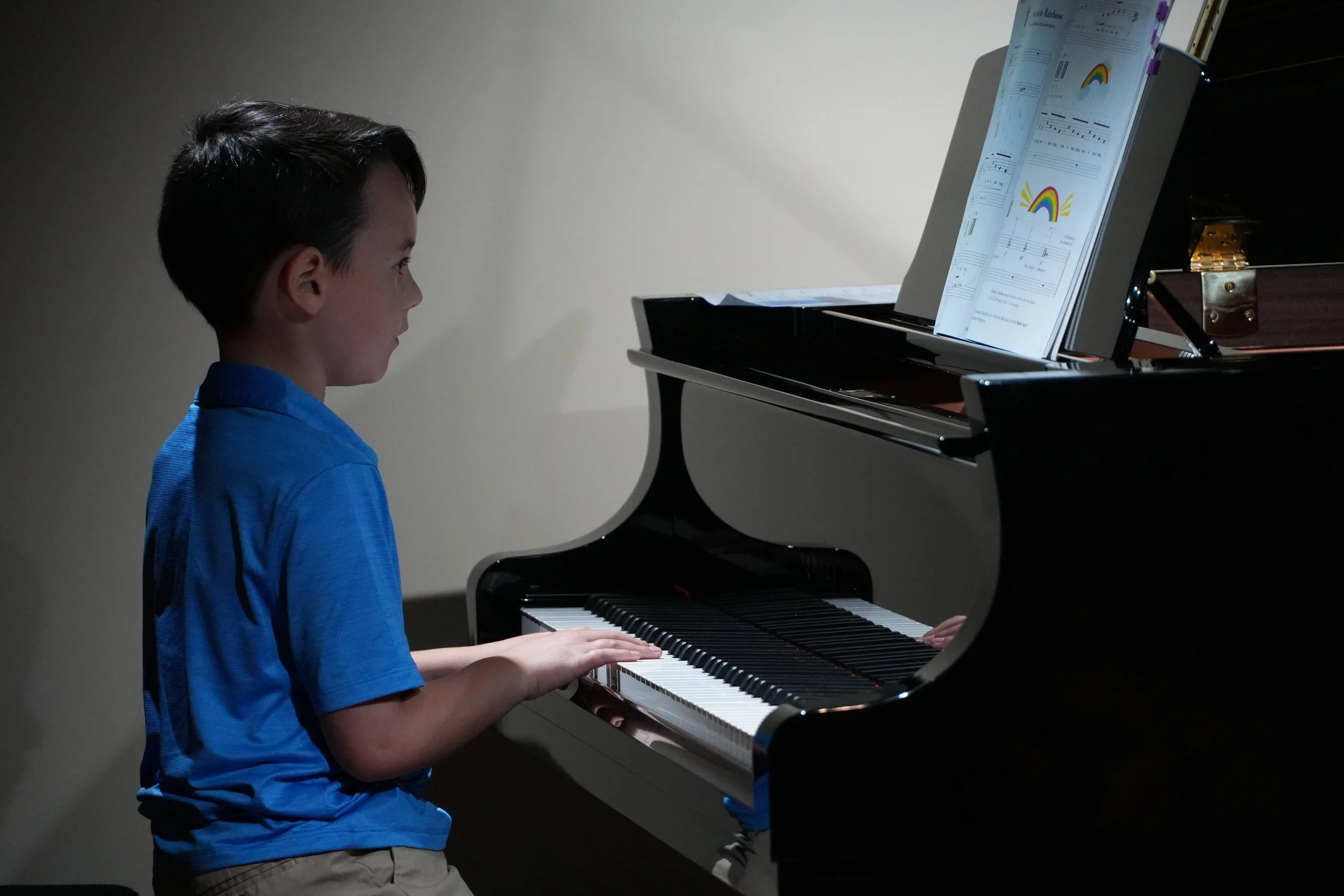 A young boy wearing a blue shirt playing a grand piano with sheet music and drawings on his music stand.