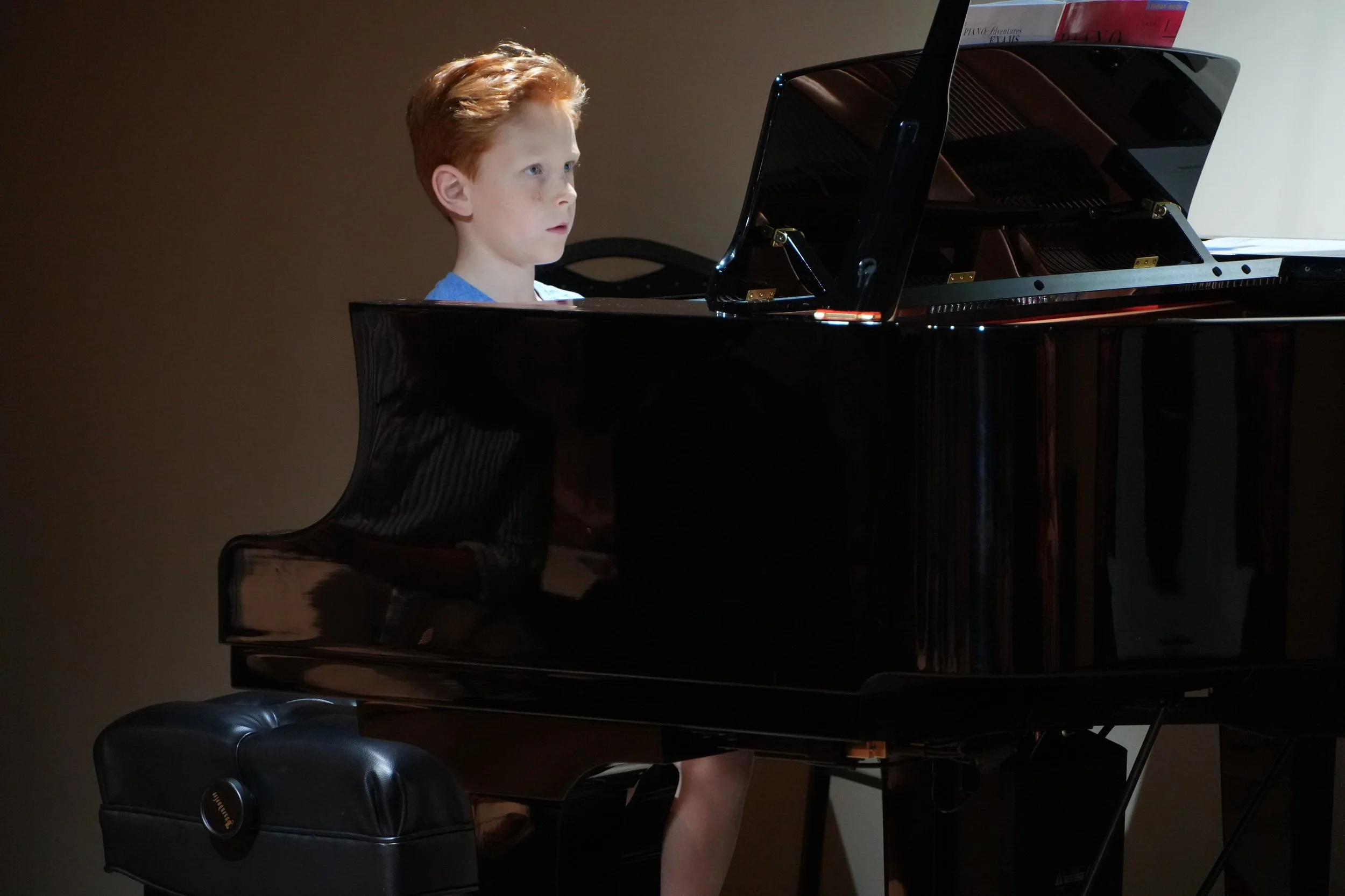 A young boy with red hair and a blue shirt is playing a grand piano with sheet music on top, in a room with a plain wall background.