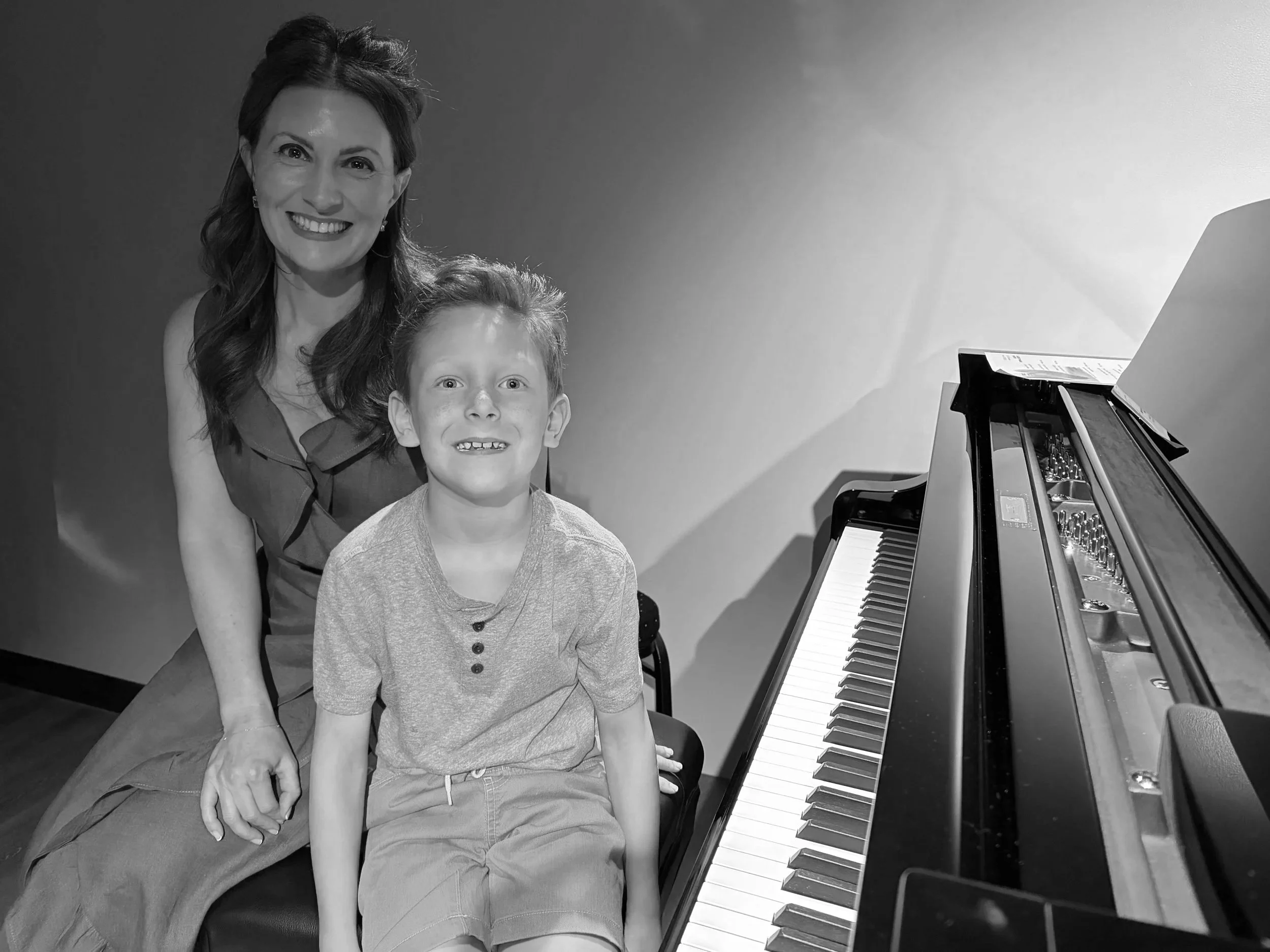A woman and a young boy pose together next to a grand piano. The woman is smiling and has dark, styled hair, and the boy is sitting with a happy expression, both indoors.