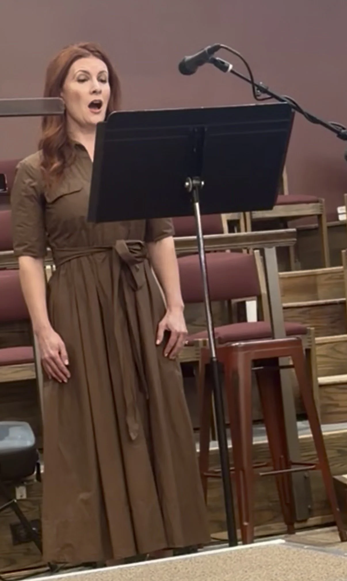 A woman singing or speaking at a microphone stand, standing in a room with stacked chairs in the background.