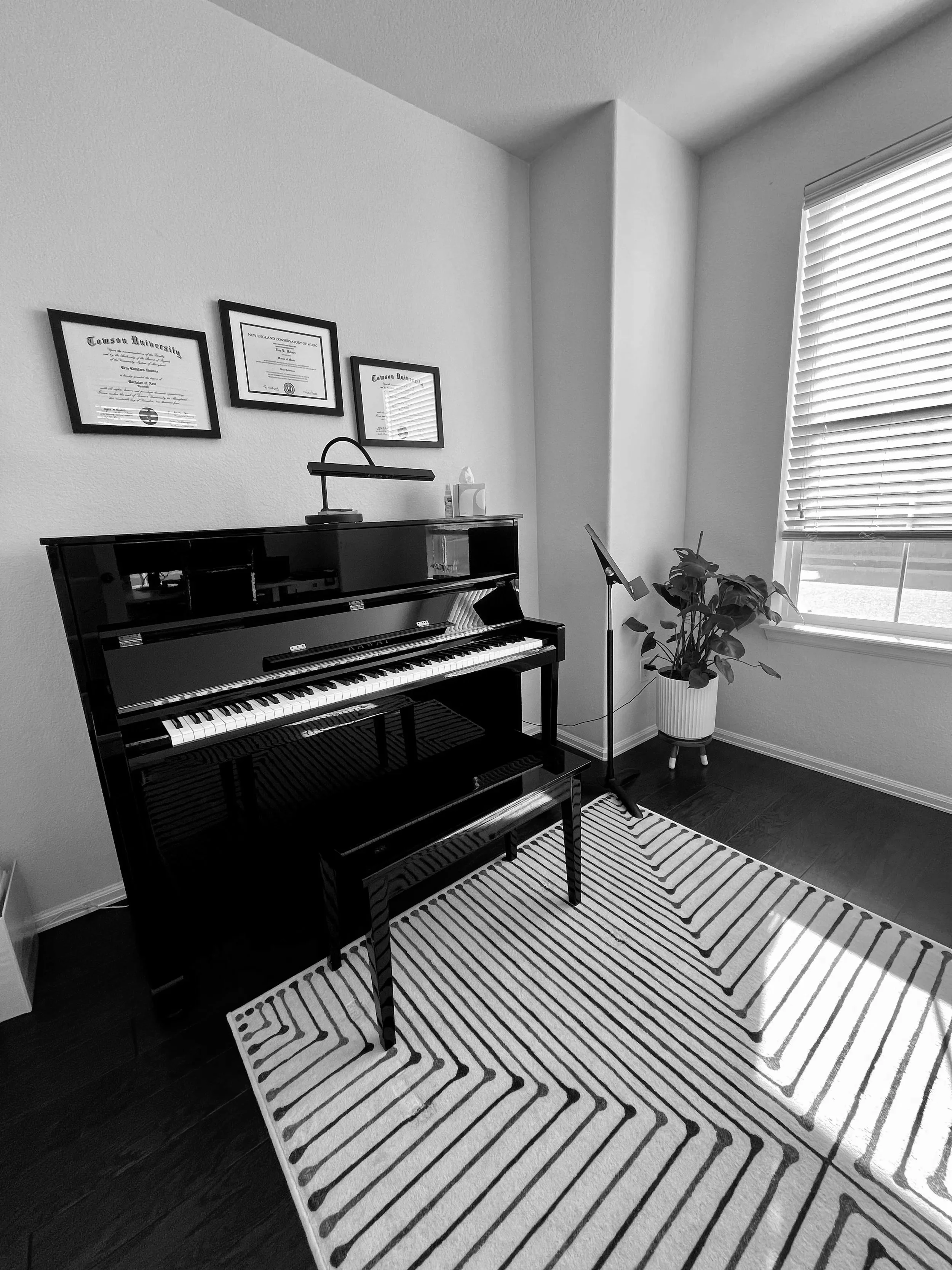 A black upright piano with a music stand and a modern table lamp on top, situated in a room with a window that has horizontal blinds, a potted plant, a guitar stand, framed diplomas or certificates on the wall, a striped rug on dark wooden floor, and a tissue box on the piano.