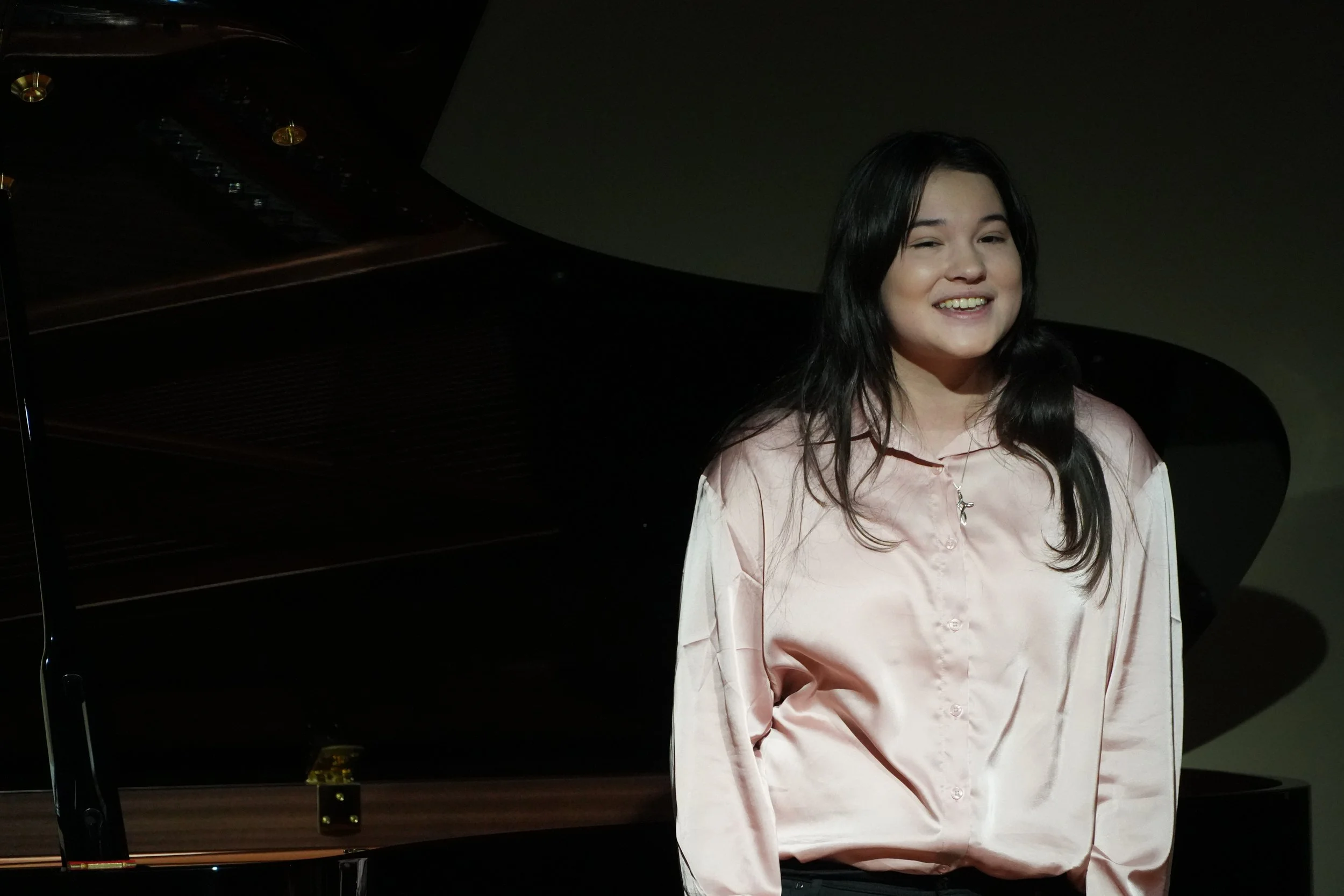 A young woman with long dark hair and a light pink satin shirt smiling in front of a black grand piano.