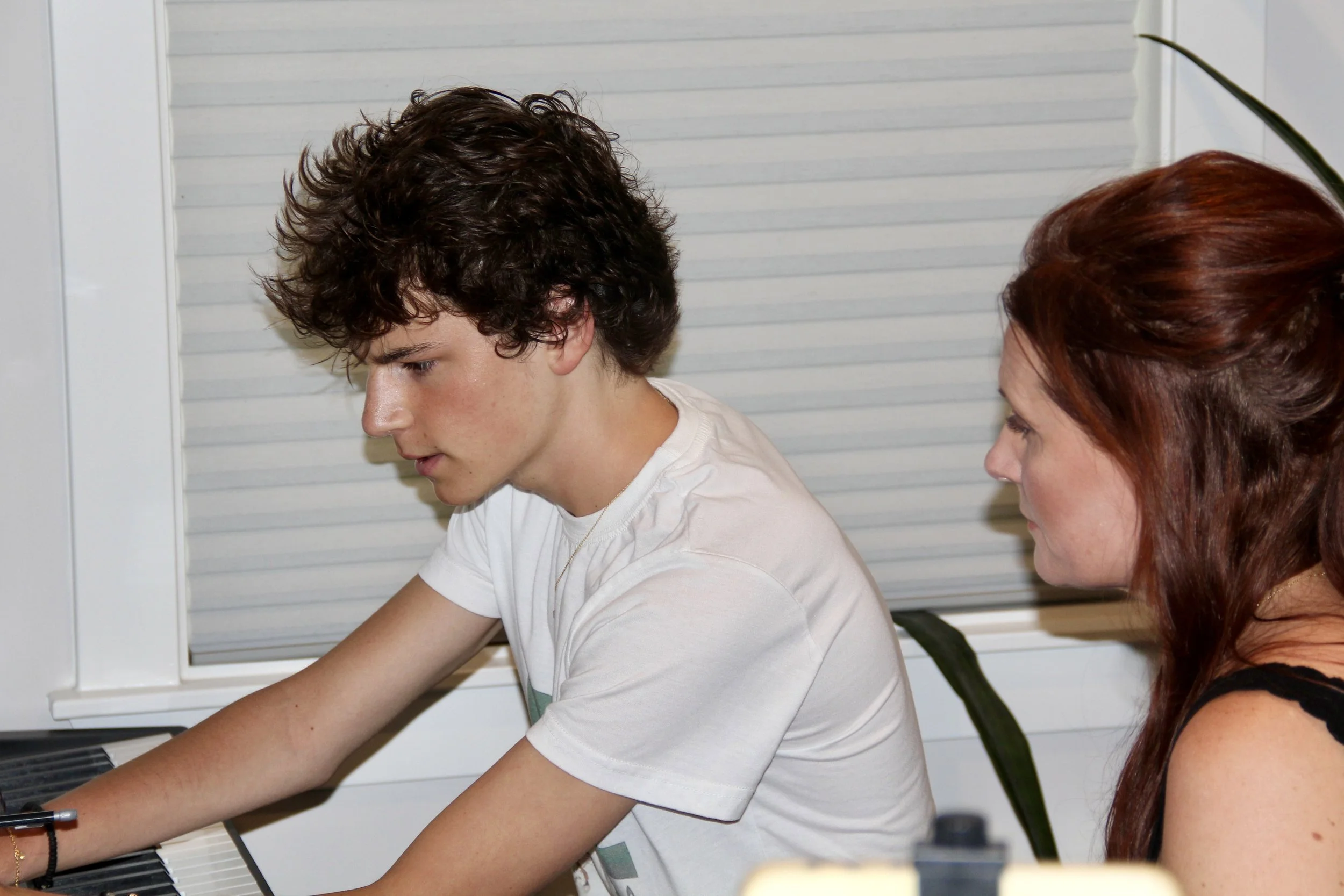A young man with curly brown hair playing a piano, focused on the keys, alongside a woman with long reddish hair.