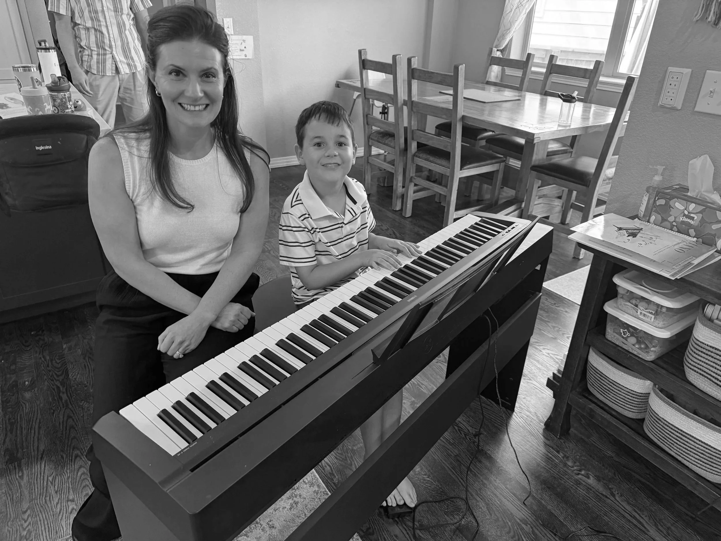 A woman and a young boy sitting at a digital piano in a dining room, smiling at the camera. The woman has long hair and is wearing a sleeveless top. The boy is wearing a striped polo shirt. There are chairs around the table, and a window with curtains in the background.