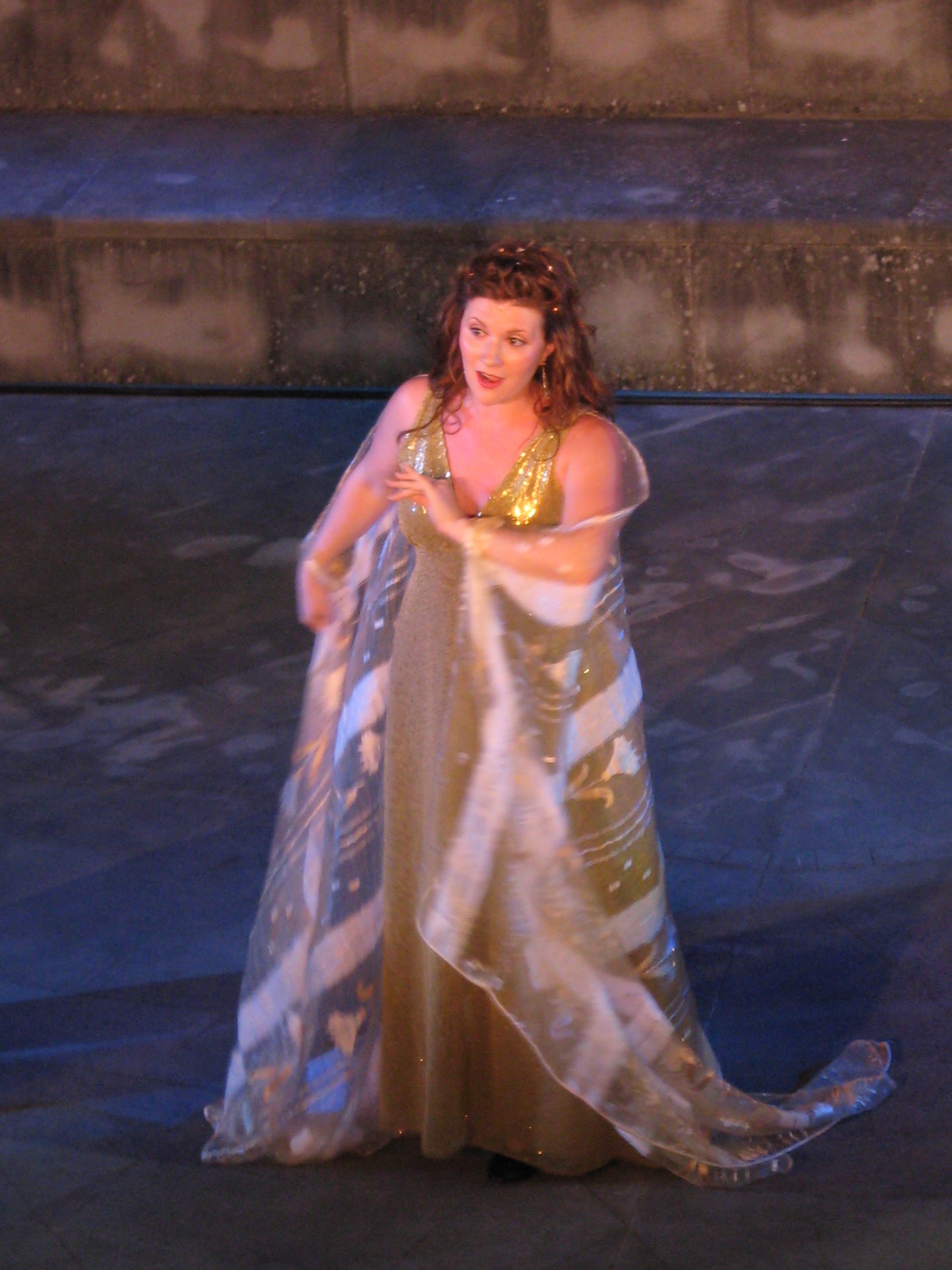 A woman with red curly hair wearing a gold and cream-colored dress, standing under a bridge or overpass at night.