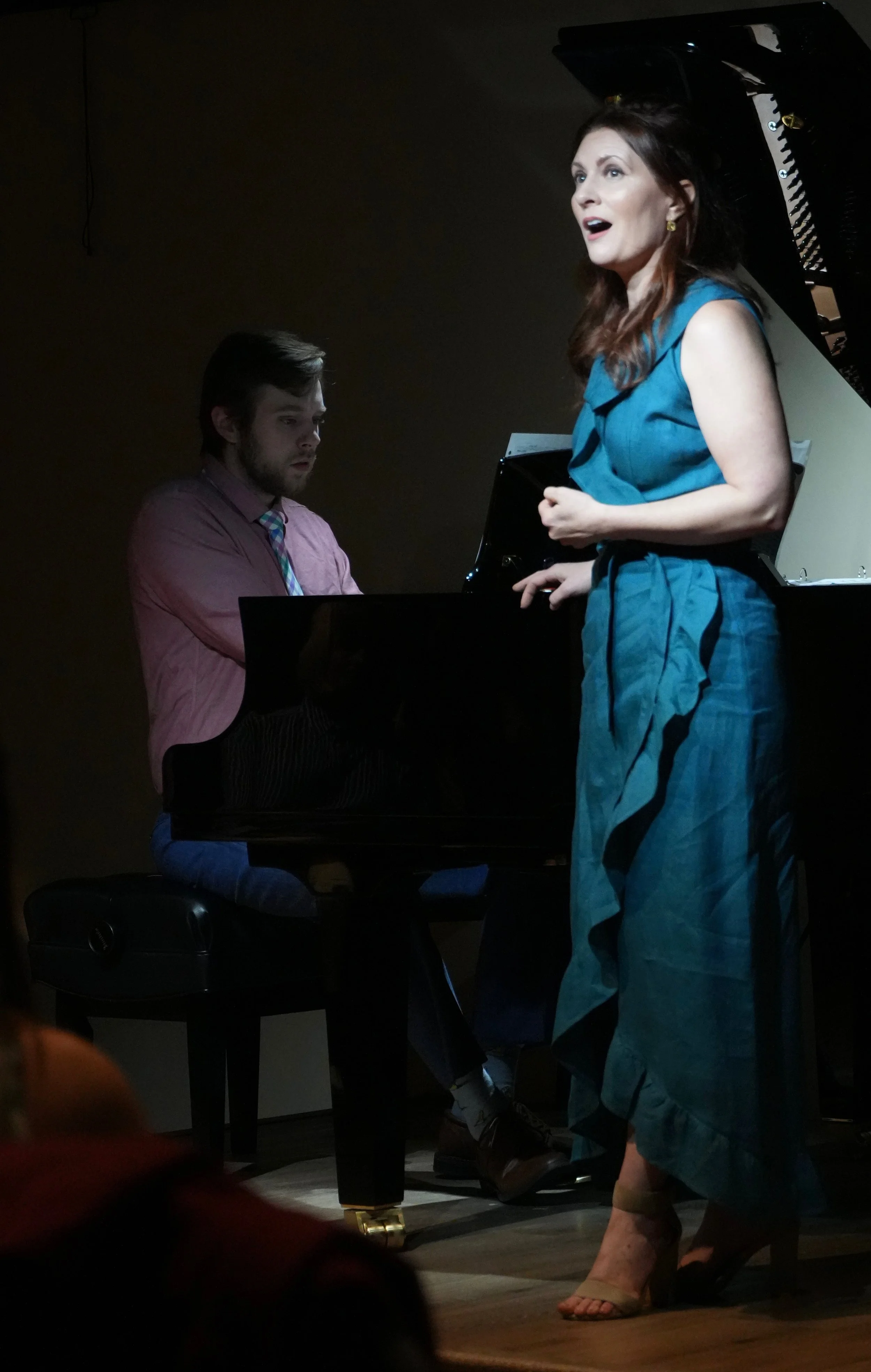A woman singing passionately at a grand piano with a man playing the piano beside her. The woman is wearing a blue dress and high heels, with her hands clasped at her waist. The scene appears to be a performance or concert.