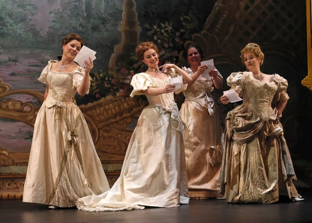 Four women dressed in elegant, vintage-style gowns stand on a stage with ornate, classical backdrop, holding papers, possibly performing in a theatrical production or operatic scene.