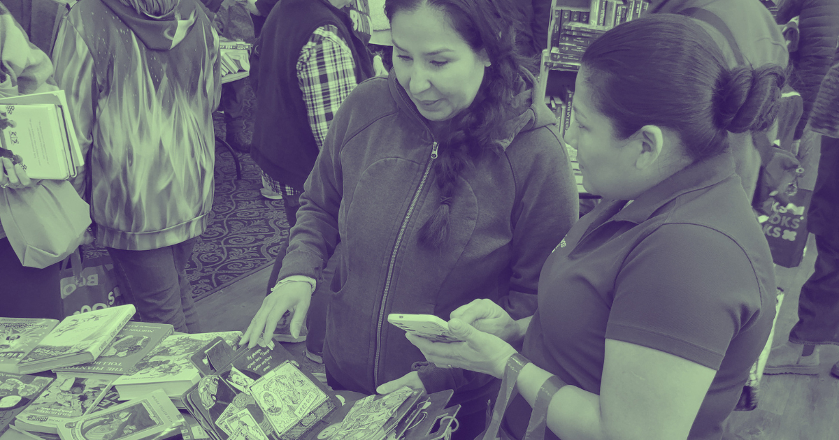 Two women browsing books at a table in a busy bookstore or event, with other people in the background.