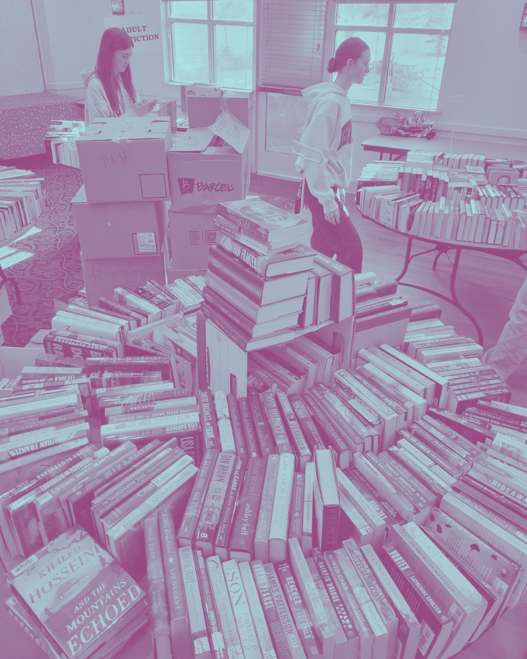 Two women browsing books inside a bookstore with tables and boxes filled with books.