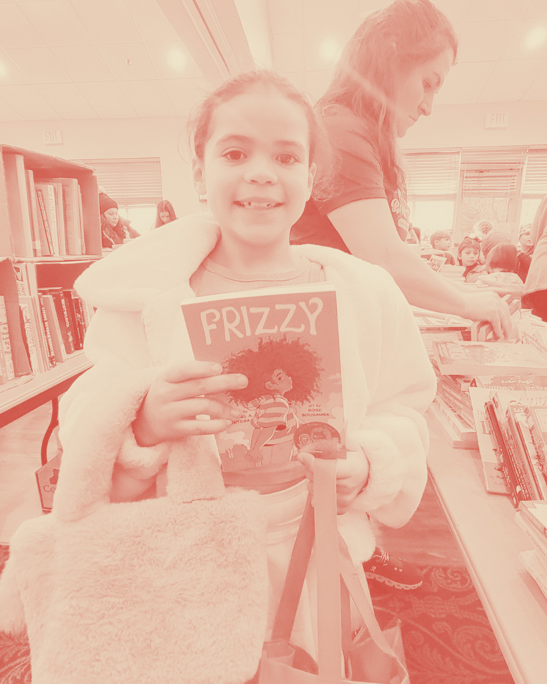 A young girl holding a book titled 'Prizzy' and smiling at the camera, with people browsing bookshelves in a library background.