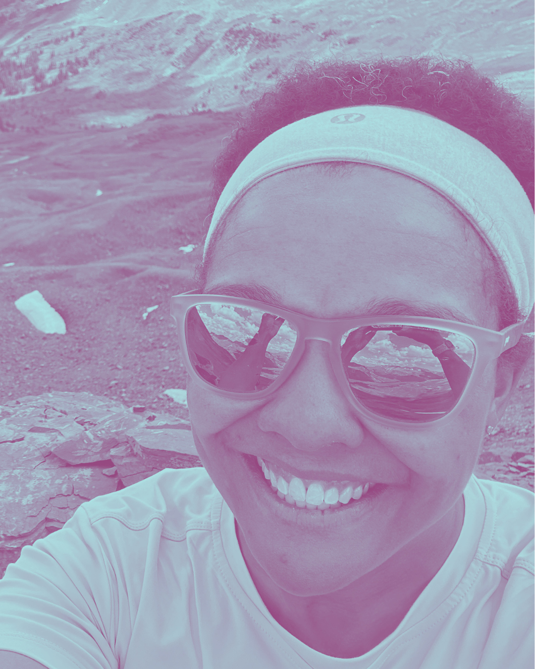 A woman smiling, wearing sunglasses and a headband, taking a selfie outdoors near rocky terrain with mountains and sky reflected in her sunglasses.