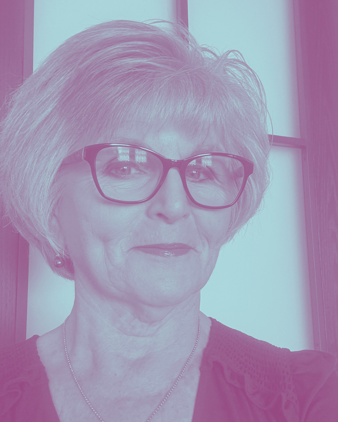 Portrait of an elderly woman with gray hair, wearing glasses, earrings, a necklace, and a dark blouse, standing indoors near a window with a wooden frame.