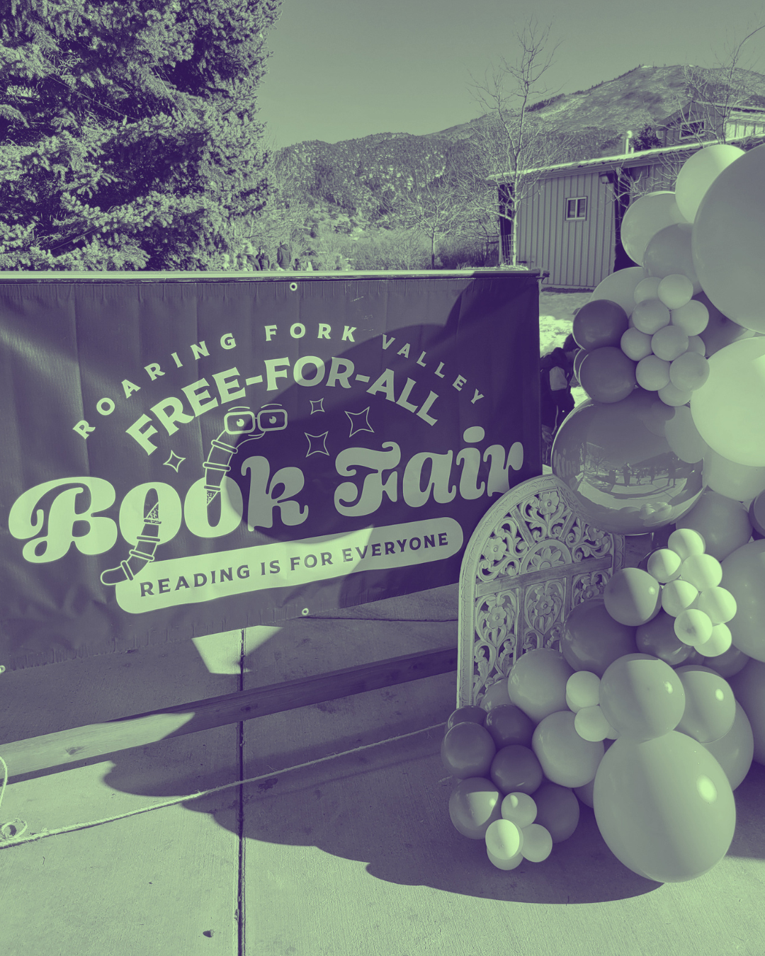 A banner with the text 'Roaring Fork Valley Free-for-All Book Fair Reading is for Everyone' is displayed outdoors next to a balloon arrangement with various sized balloons in light and dark shades. In the background, there are trees, mountains, and a building, suggesting a scenic outdoor setting.