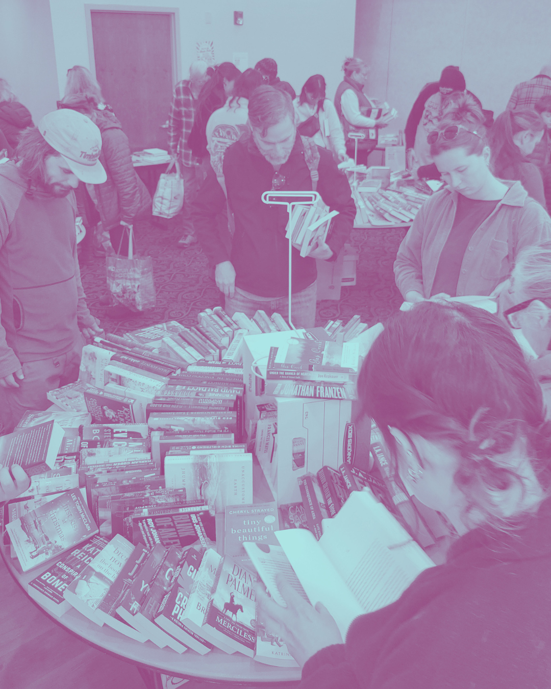 People browsing and reading books at a book sale or event with multiple tables filled with books in a room with light-colored walls.