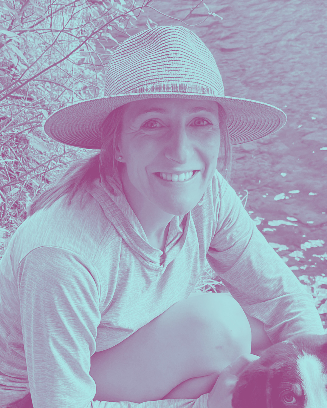 A woman smiling while sitting by a body of water, wearing a wide-brimmed hat and a long-sleeve shirt, with a dog partially visible in the foreground.